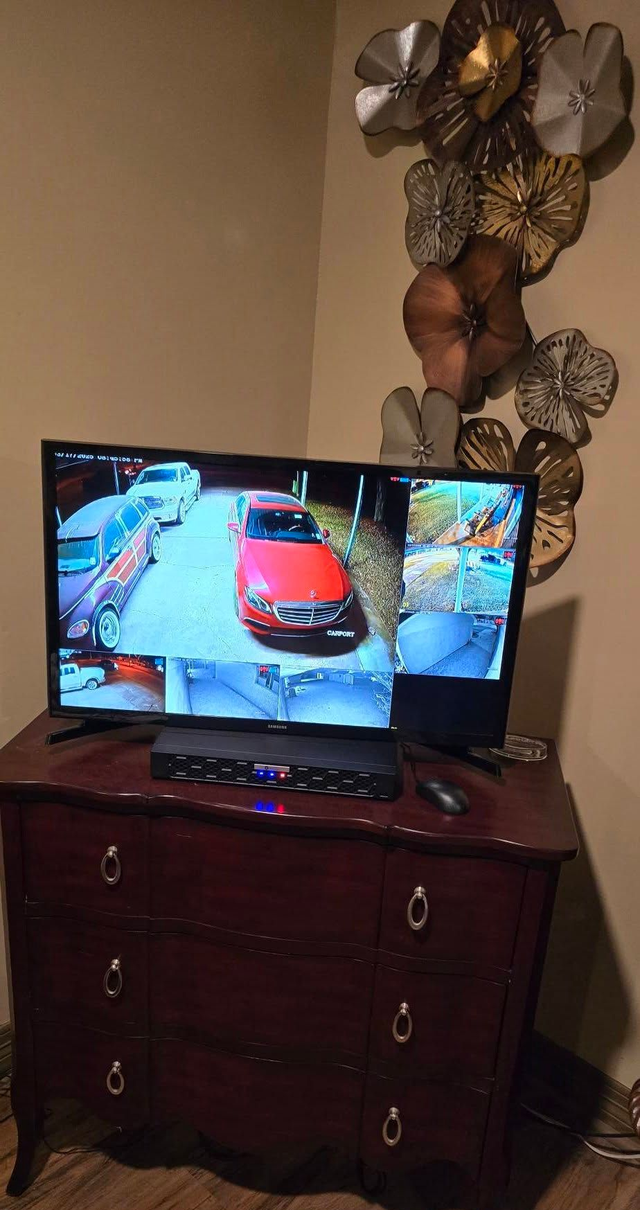 TV on a wooden dresser displaying surveillance camera views of a driveway with cars. Decorative wall art on the right.