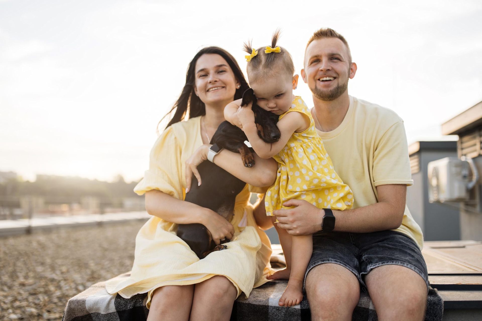 A family wearing yellow outfits sits together outdoors at sunset, holding a small dark dog.