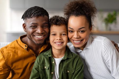 Three smiling people pose together indoors, wearing casual clothing in earthy tones, against a soft-focus background.