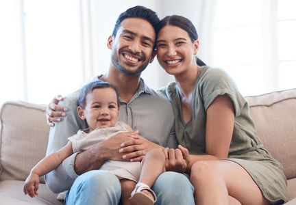 A smiling family of three sitting together on a beige couch in a brightly lit room.