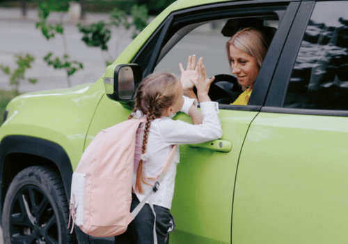 Girl with pink backpack high-fives a woman in a green car through the window.