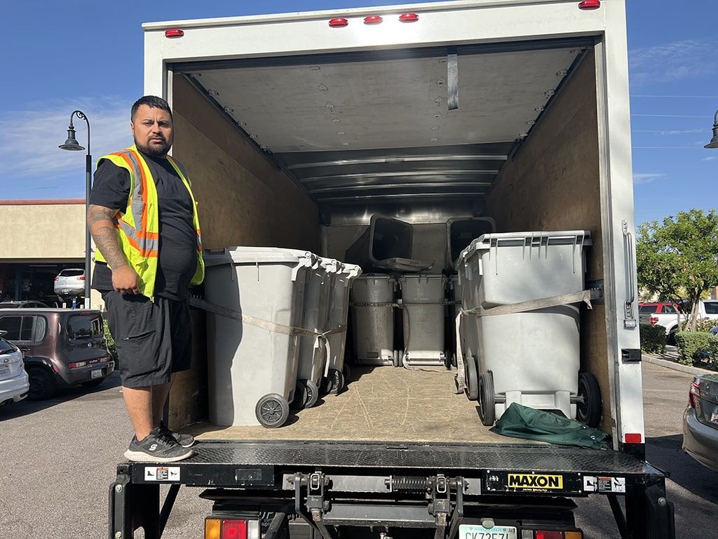 Worker standing by a moving truck loaded with trash bins and containers.