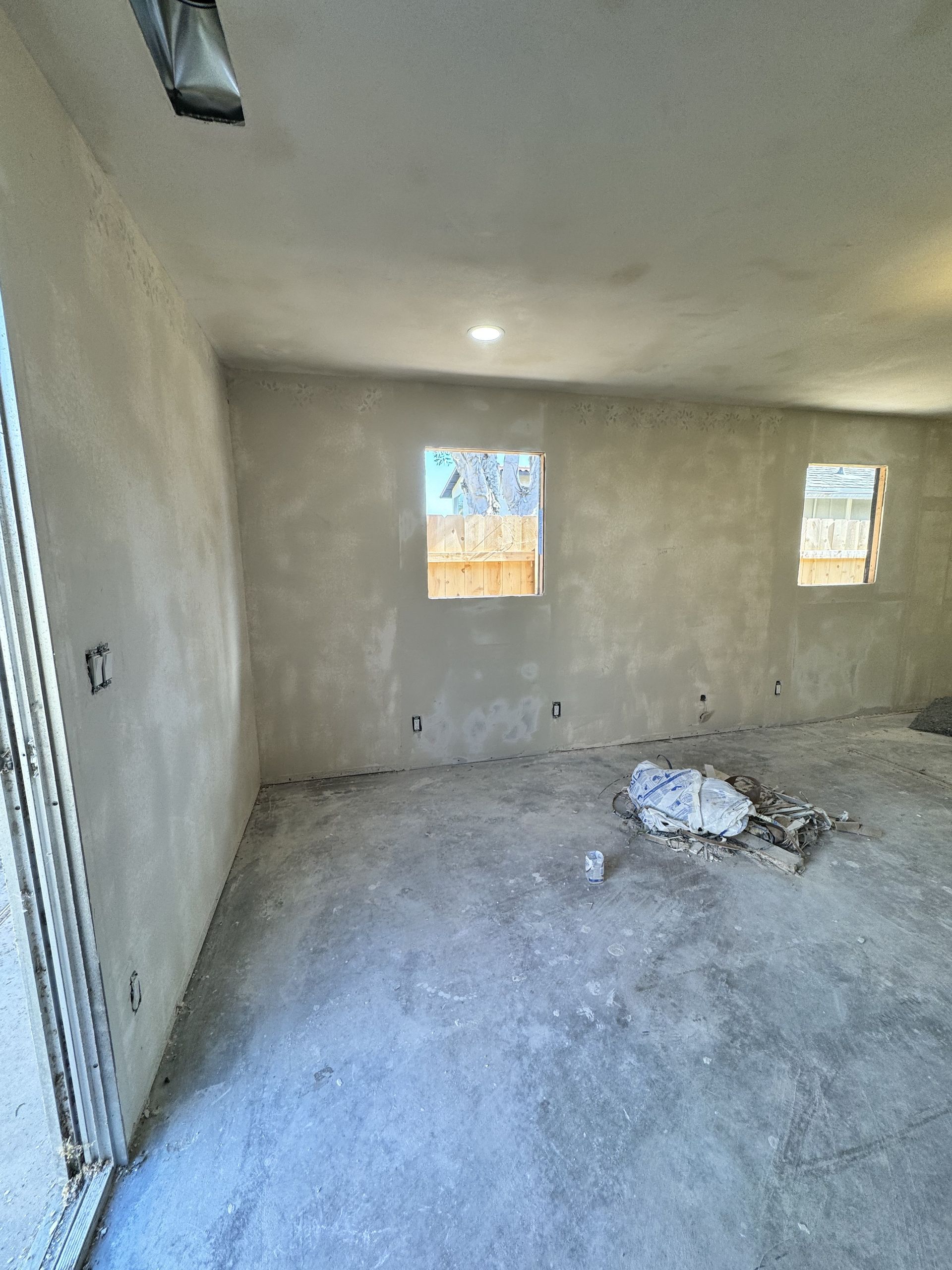 Bare unfinished room with gray walls, small windows, and debris on the concrete floor.