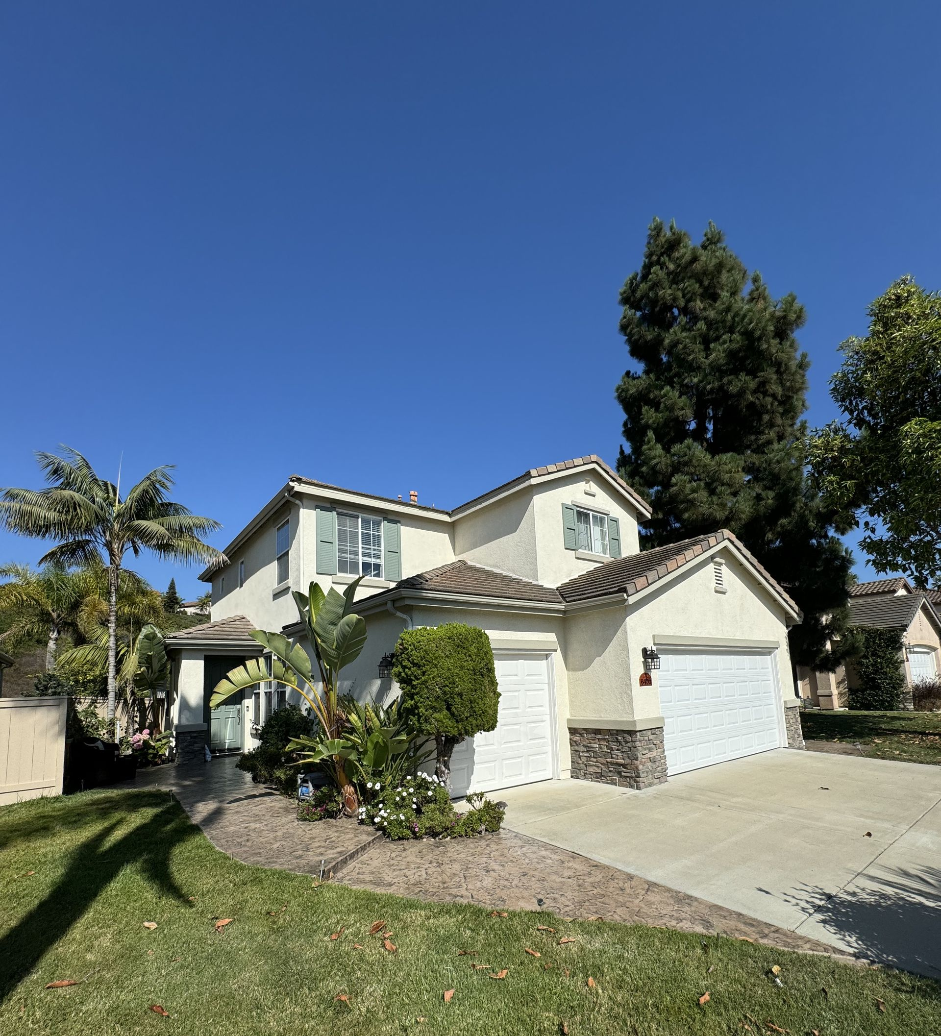 Suburban two-story white house with a driveway, front lawn, and tall trees under a clear blue sky