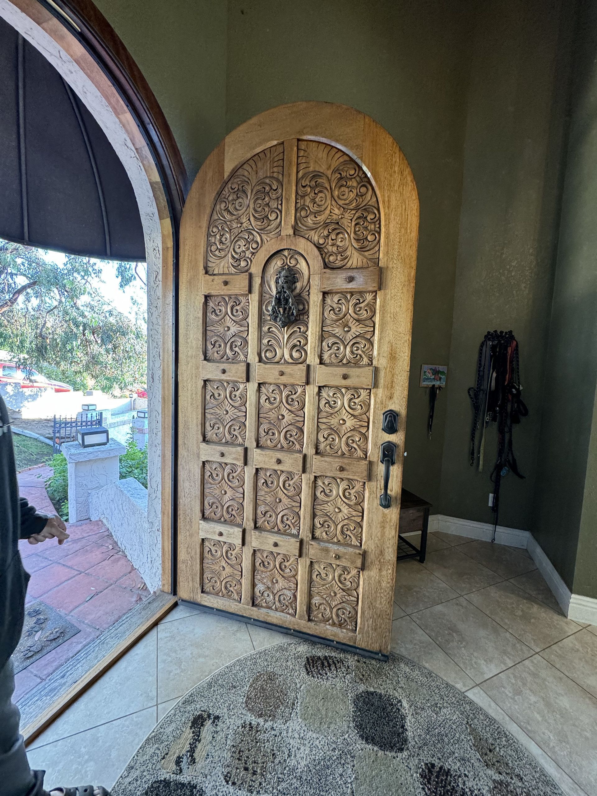 Open wooden door with ornate panels beside an arched window in a tiled entryway