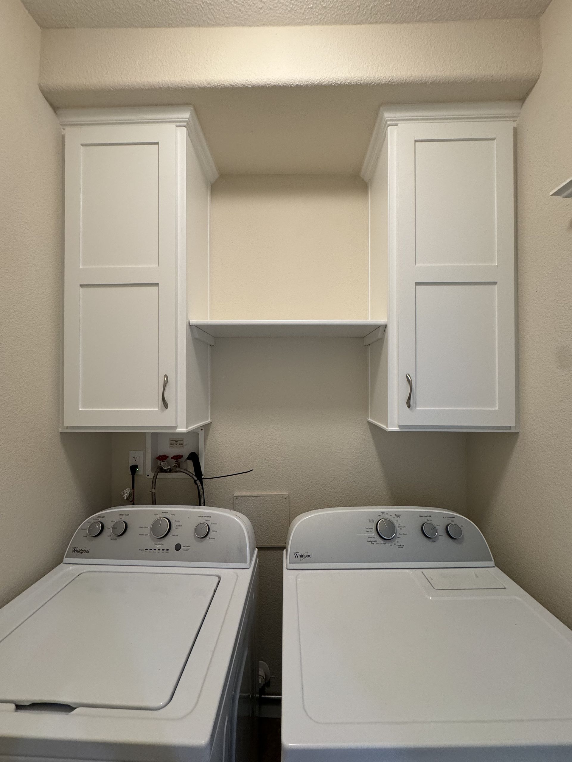 Laundry room with white washer and dryer under wall cabinets and a shelf between them