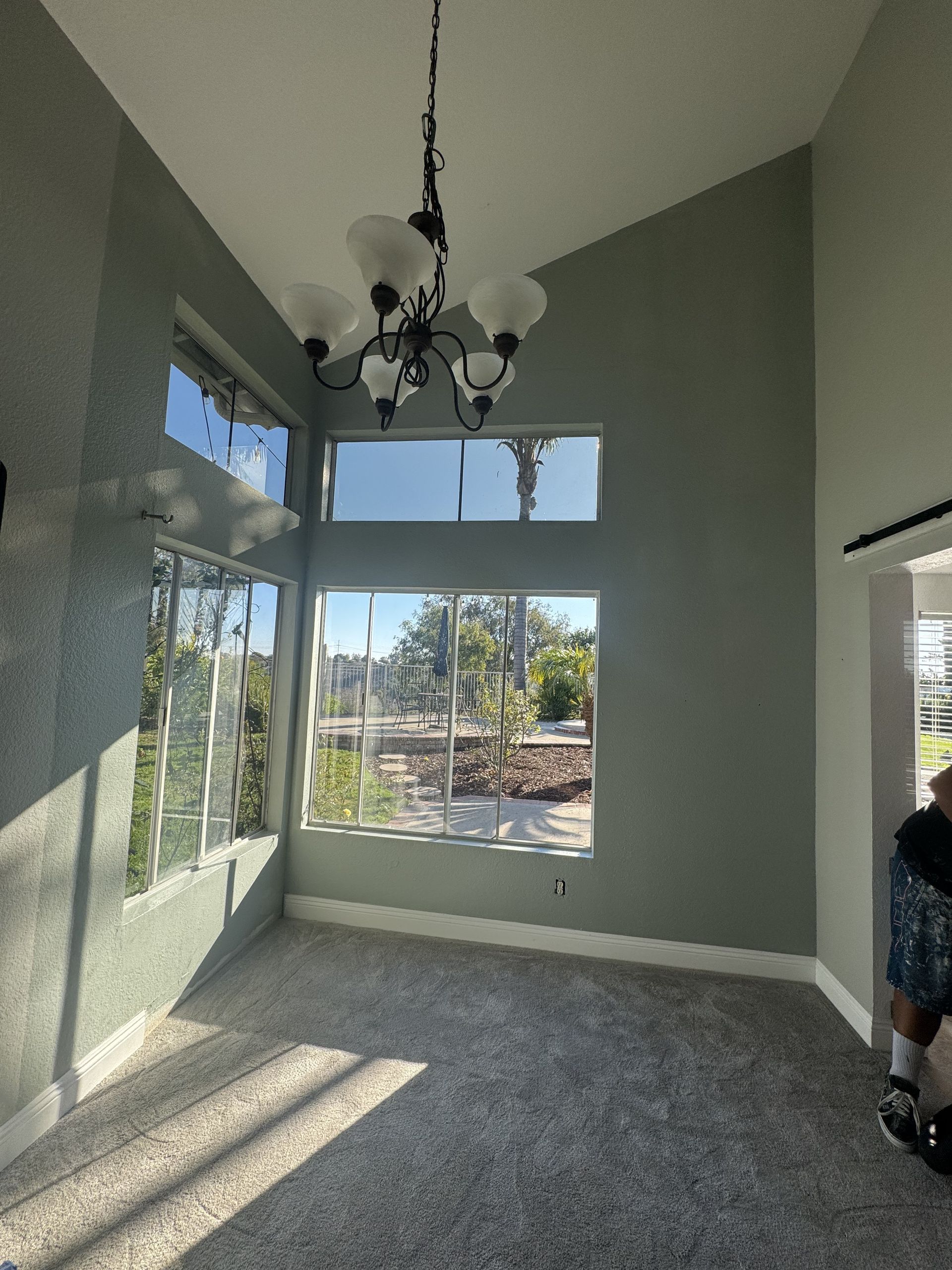 Empty modern room with gray carpet, tall windows, and a black chandelier hanging from the vaulted ceiling