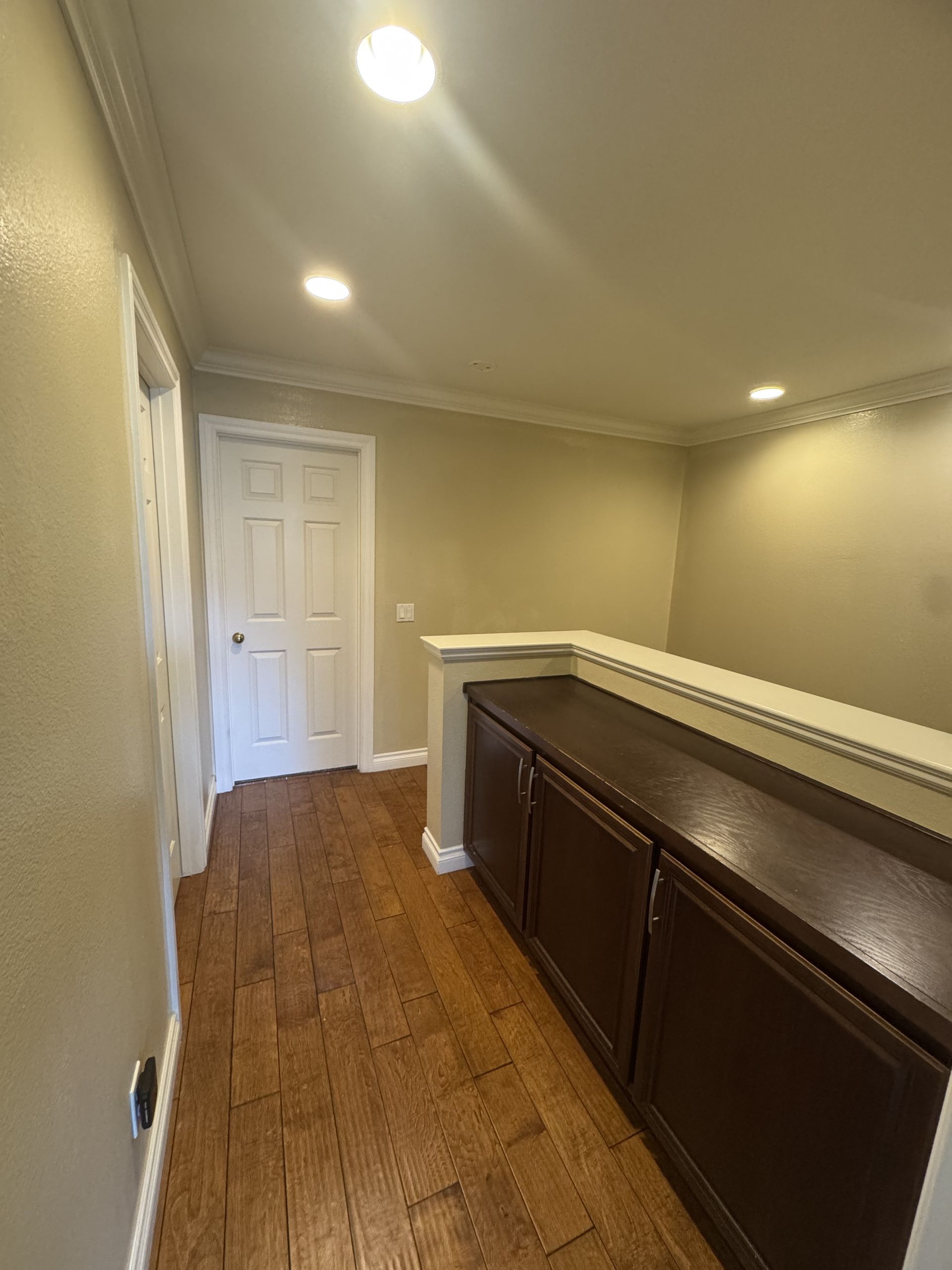 Narrow hallway with beige walls, recessed lights, white door, and dark wood cabinet railing.