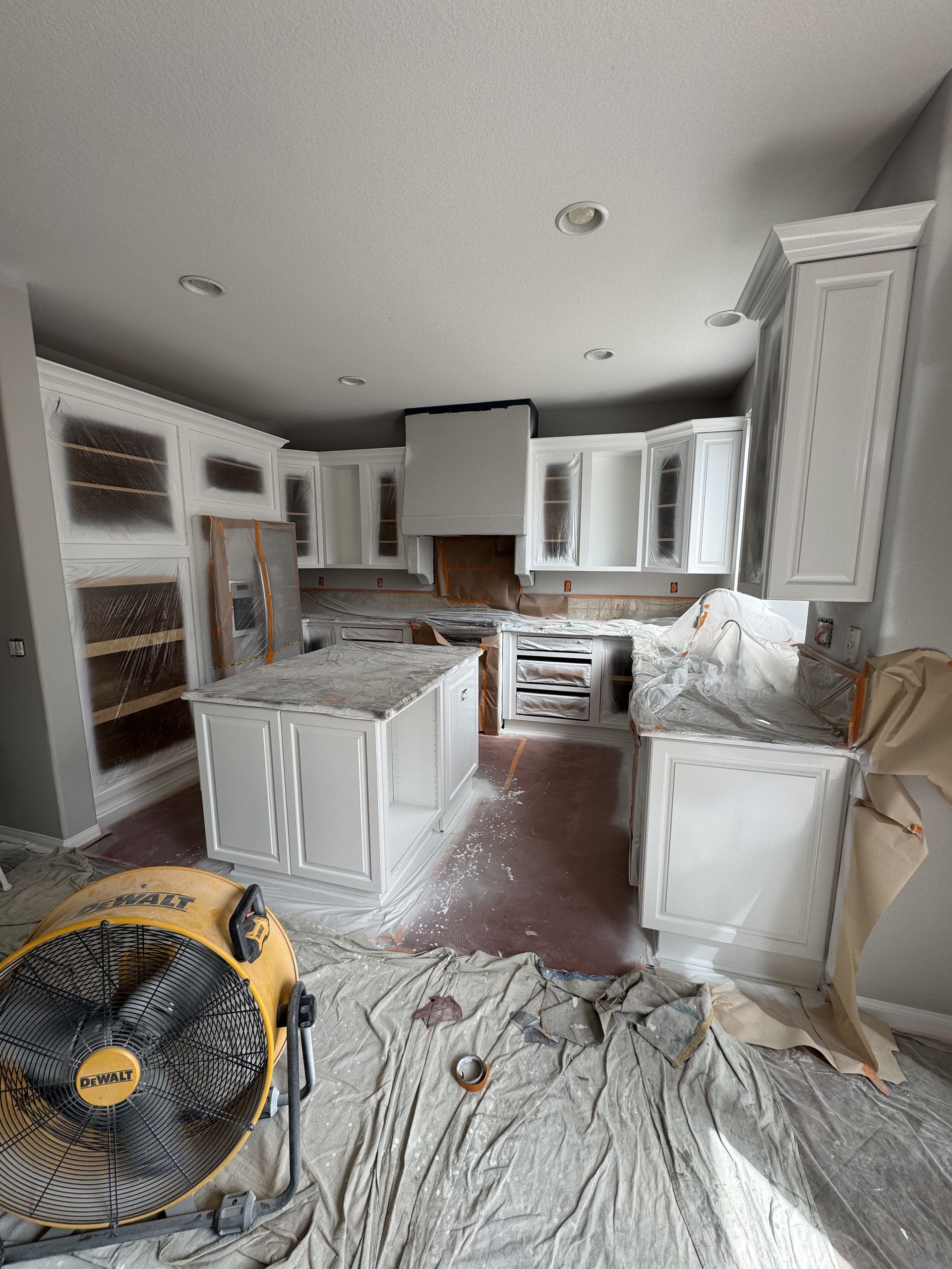 Renovating kitchen with white cabinets, exposed flooring, and covered surfaces in a bright room.