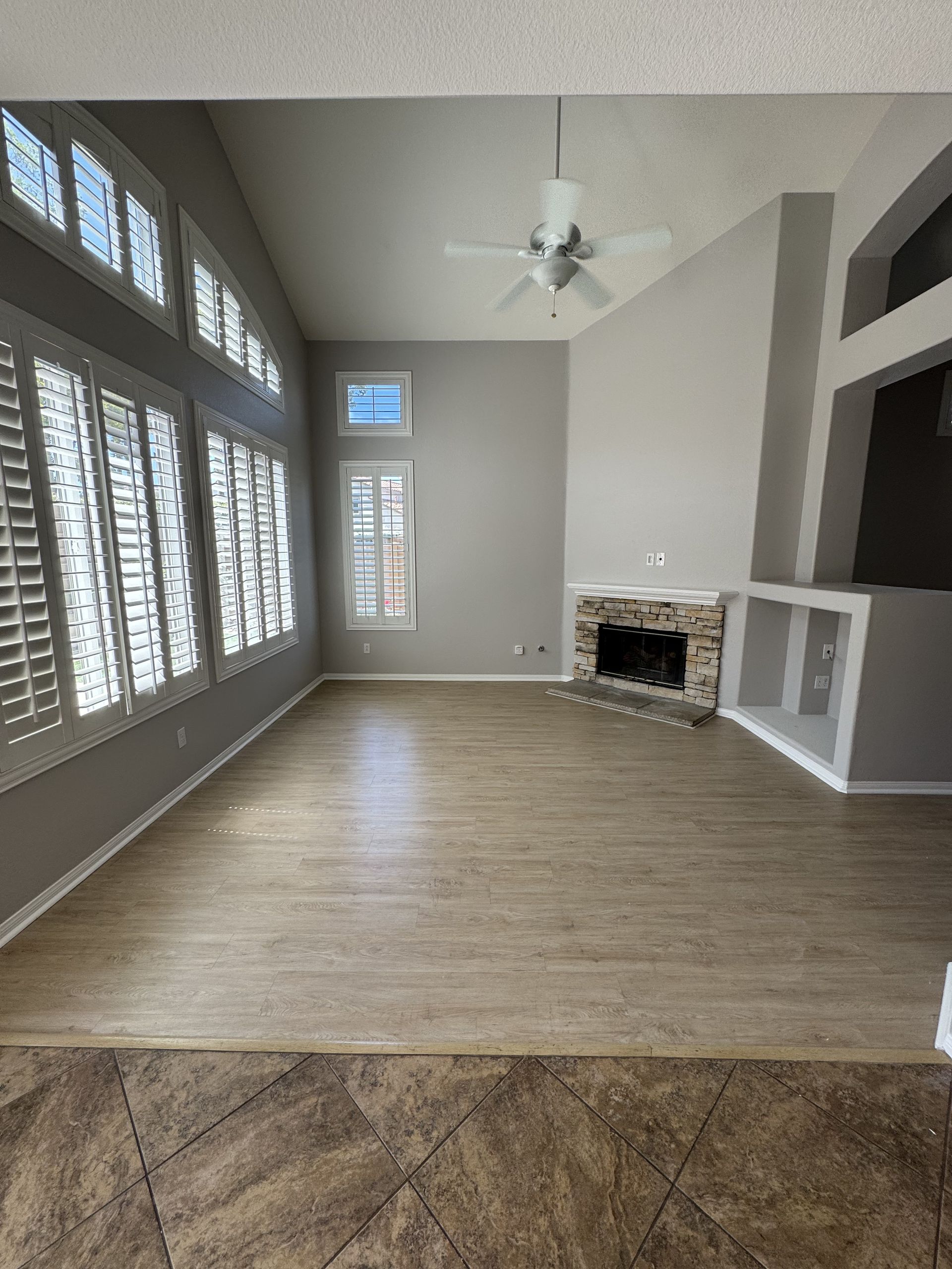 Empty living room with large windows, gray walls, ceiling fan, and a fireplace on the right