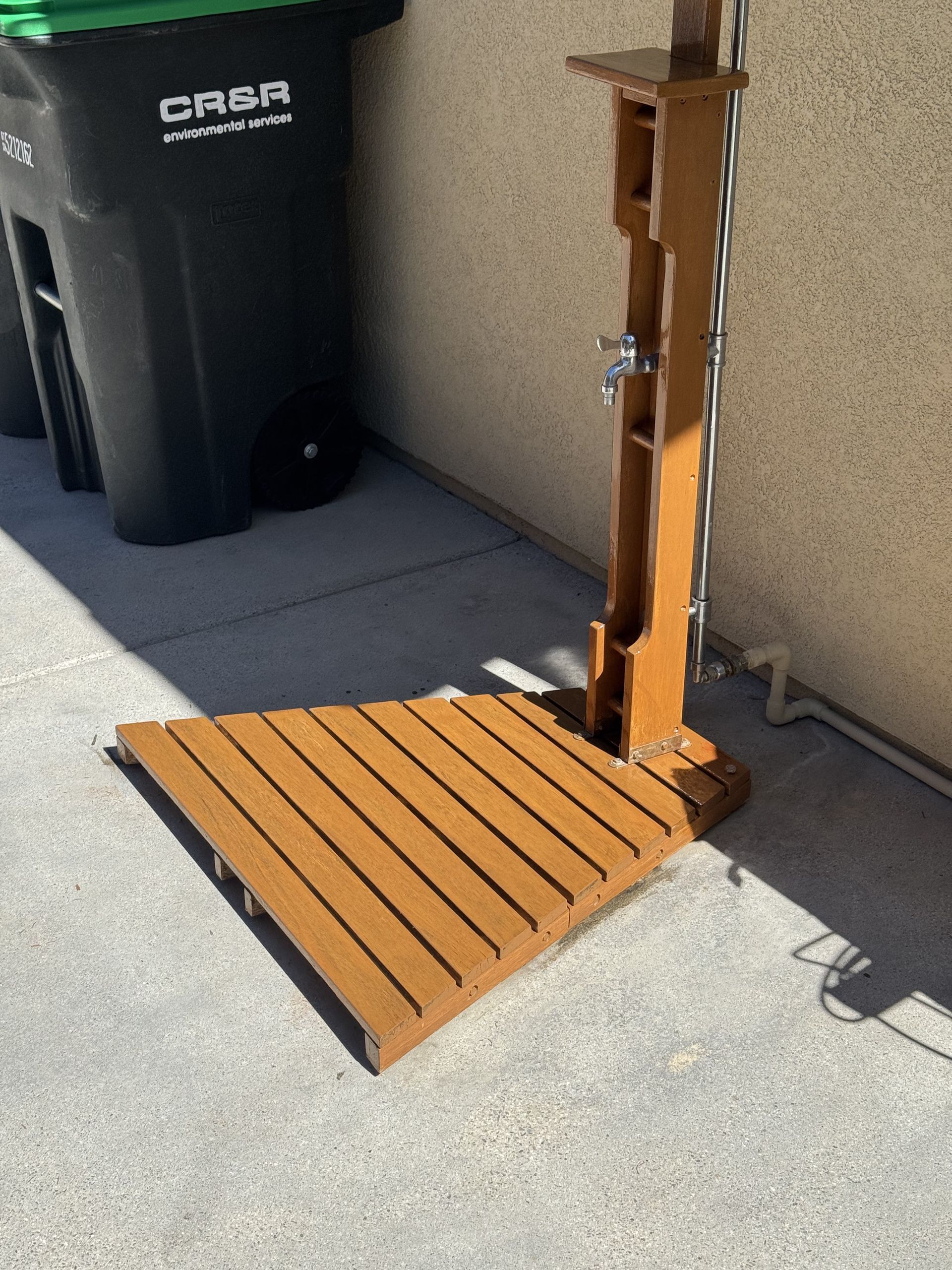 Wooden foldable ramp leaning against a wall beside a trash bin on a concrete sidewalk.