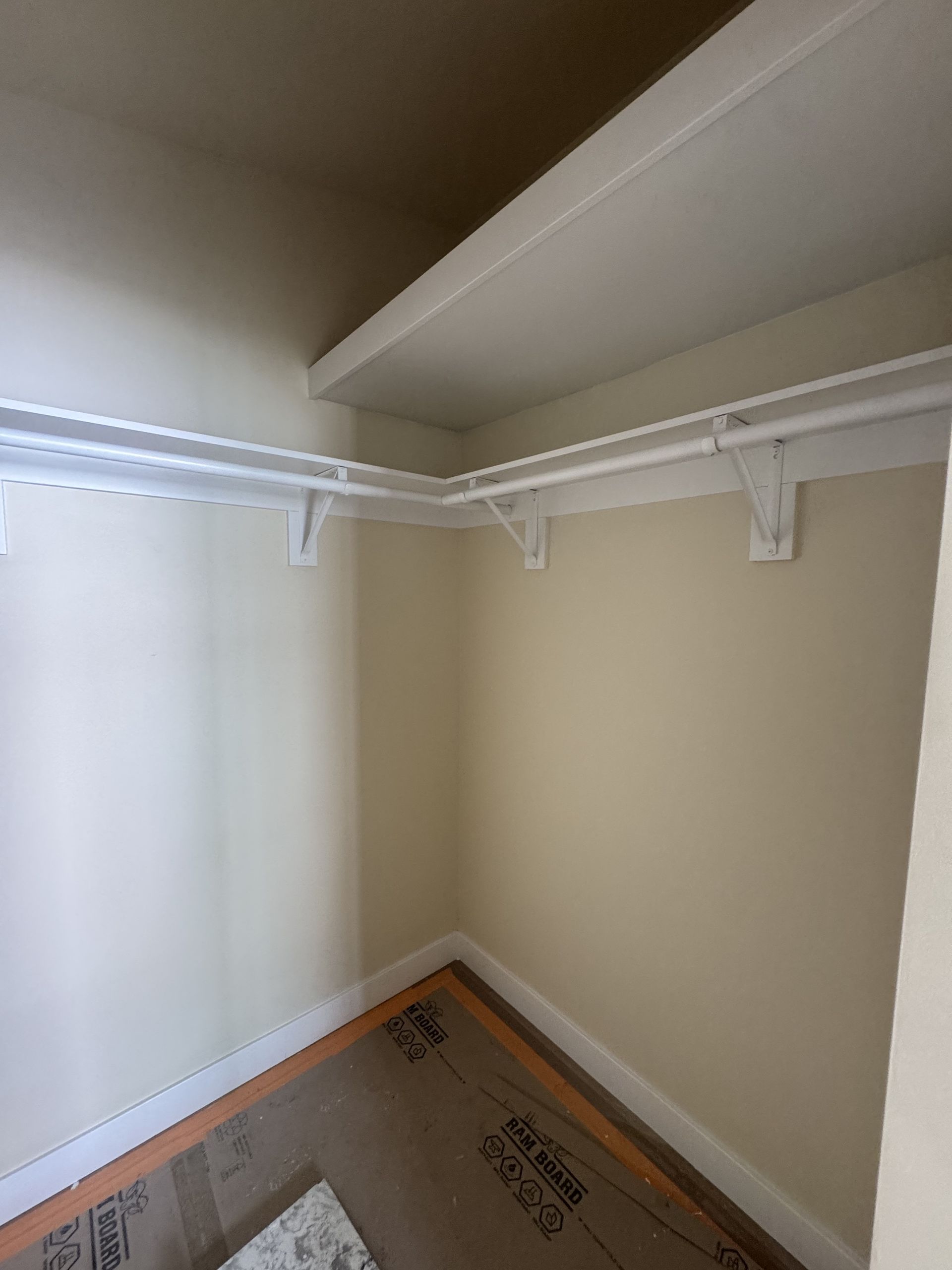 Empty closet with white shelves and hanging rod, beige walls, and a bare floor.