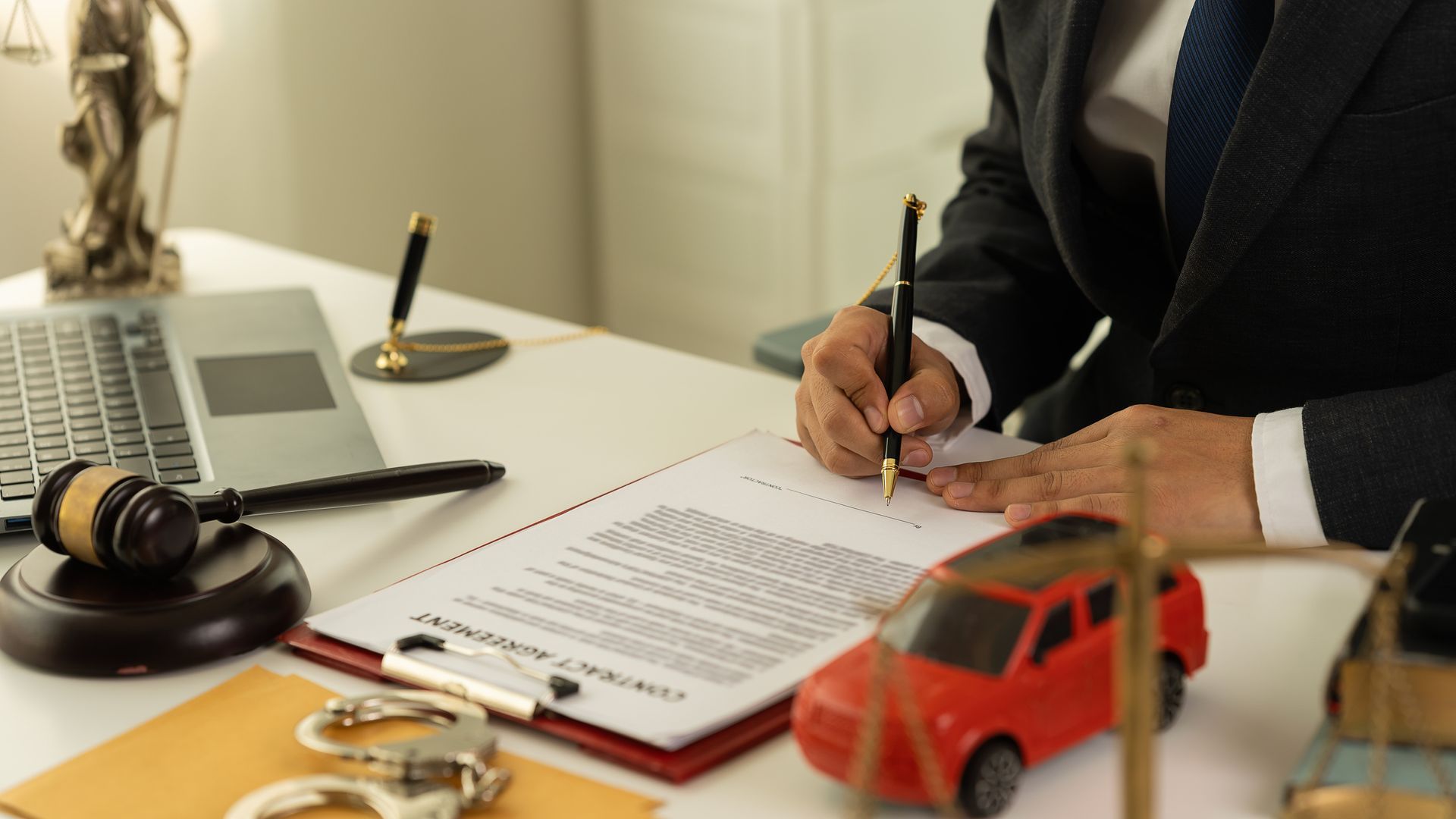 Lawyer signs contract at desk with gavel, laptop, and toy car.