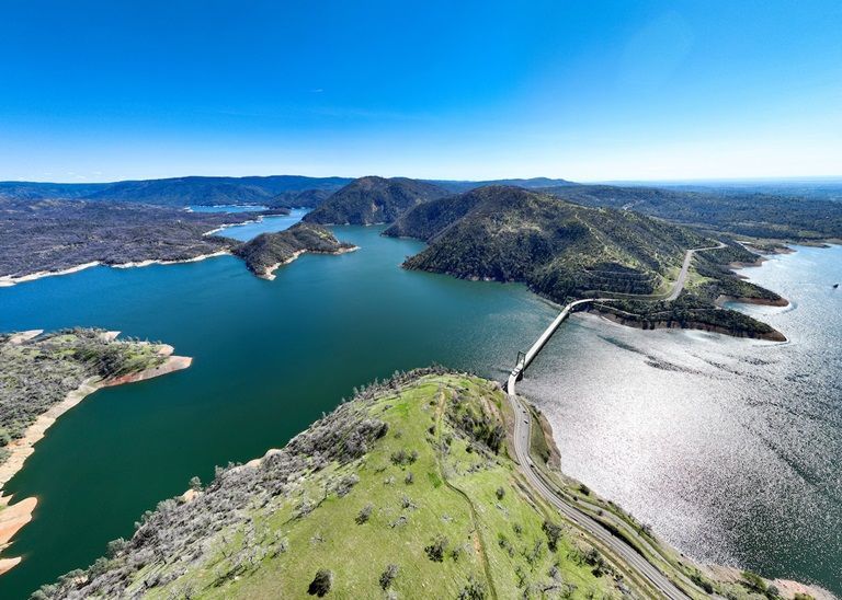 Aerial view of a blue bay with green hills, winding roads, and sunlit water along a rugged coastline