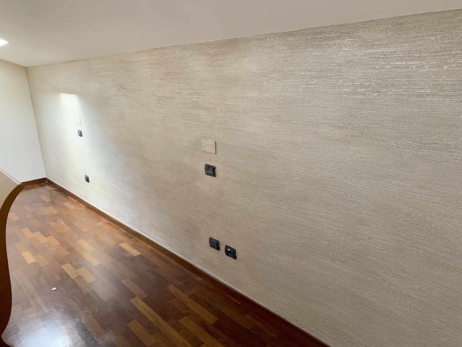 Soggiorno vuoto con pareti beige texturizzate e pavimento in legno, con prese elettriche a vista e una porta.