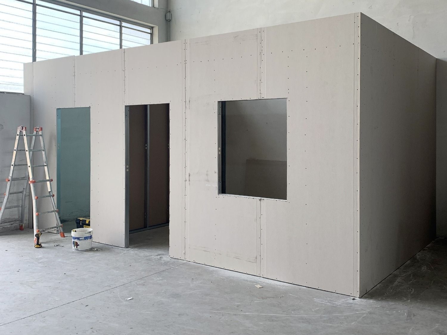 Stanza in cartongesso non rifinita in fase di costruzione, con scala e aperture per finestre all'interno di uno spazio simile a un magazzino.