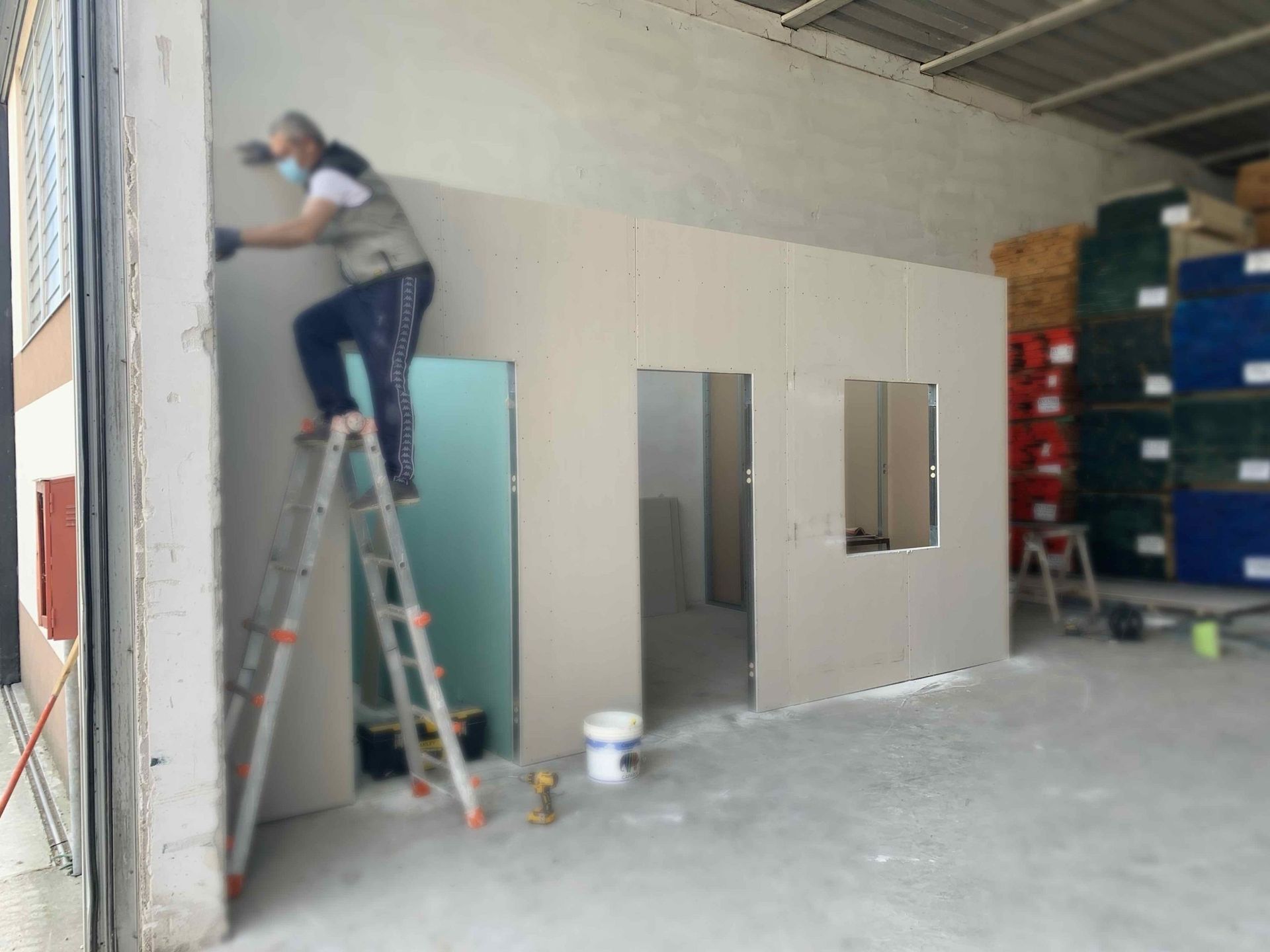Un operaio su una scala dipinge la parete di una piccola stanza all'interno di un magazzino o di uno spazio industriale.