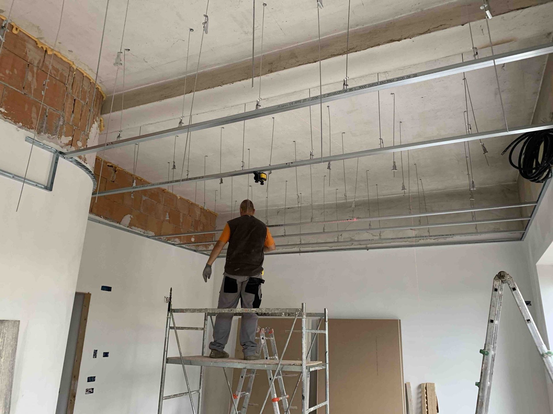 Un operaio su un'impalcatura installa un soffitto in una stanza non rifinita con mattoni e cavi elettrici a vista.