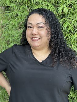Smiling person in a black shirt standing outdoors in front of green foliage