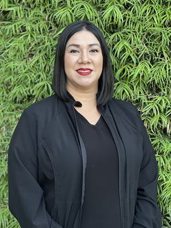 Smiling person in black jacket standing before a green leafy wall.