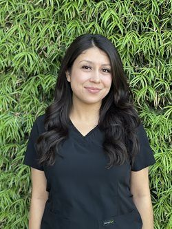 Woman in a black shirt standing in front of a green leafy wall, smiling at the camera.