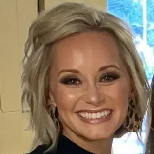 Smiling person with short blonde hair in a dark top, standing indoors by a bright window