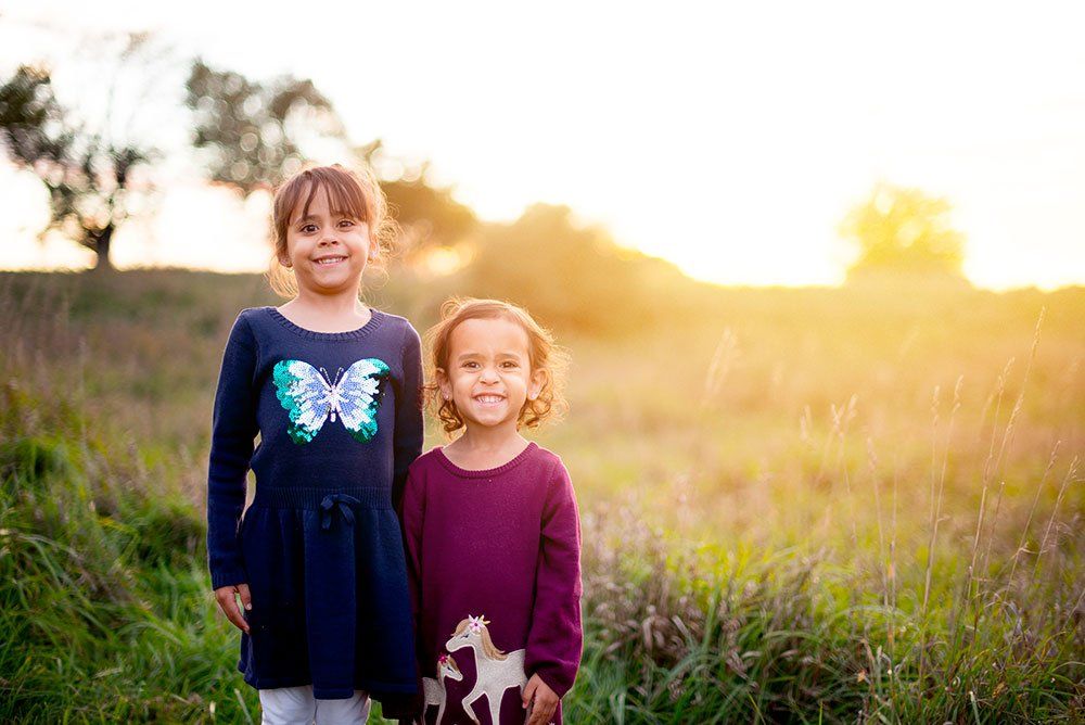 Two children smiling side by side in a grassy field during a golden sunset.