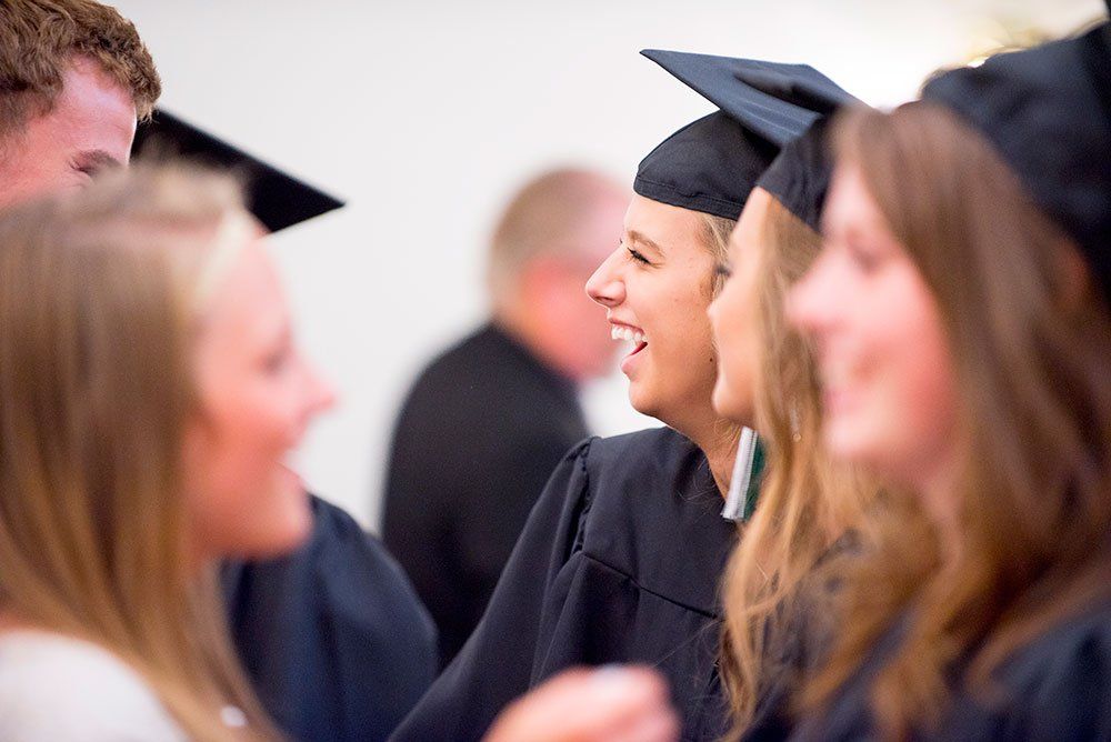 Graduates in caps and gowns laughing and chatting together in a brightly lit indoor space.