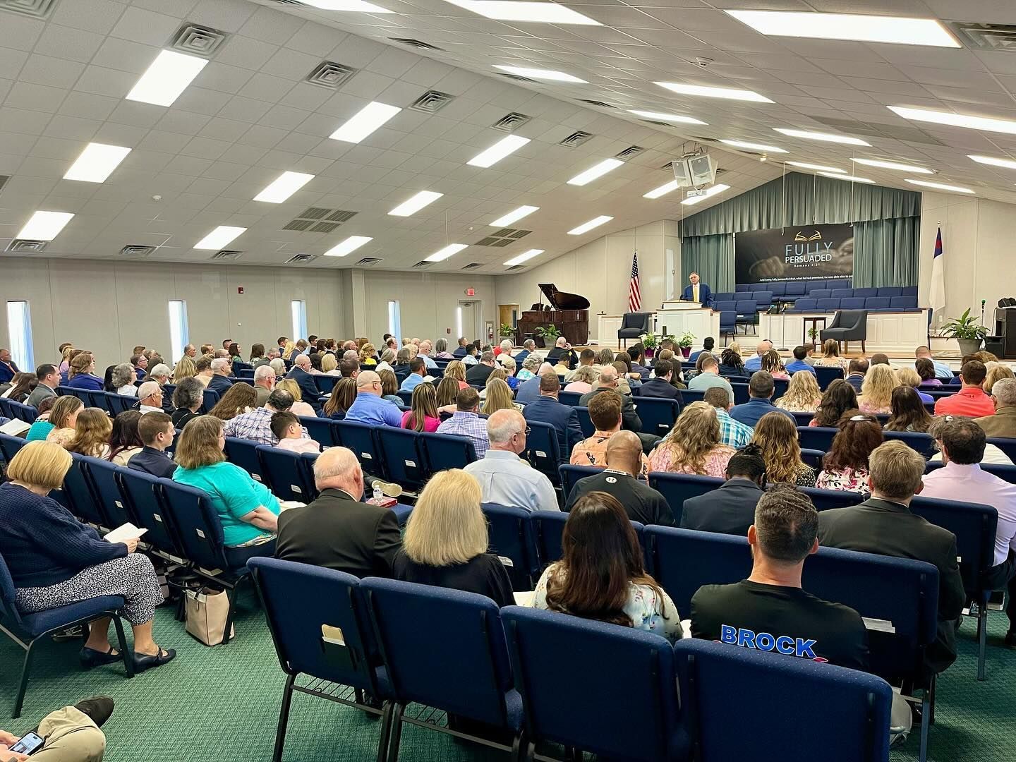 A large audience sits in chairs inside a brightly lit church sanctuary, facing a stage with a podium and American flag.