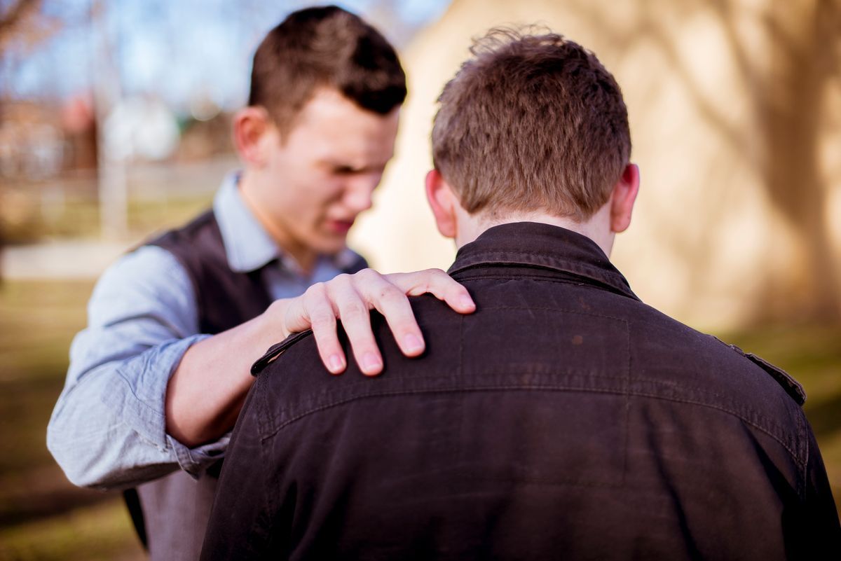 A person in a light shirt and vest gently places a hand on the shoulder of another person wearing a dark jacket.