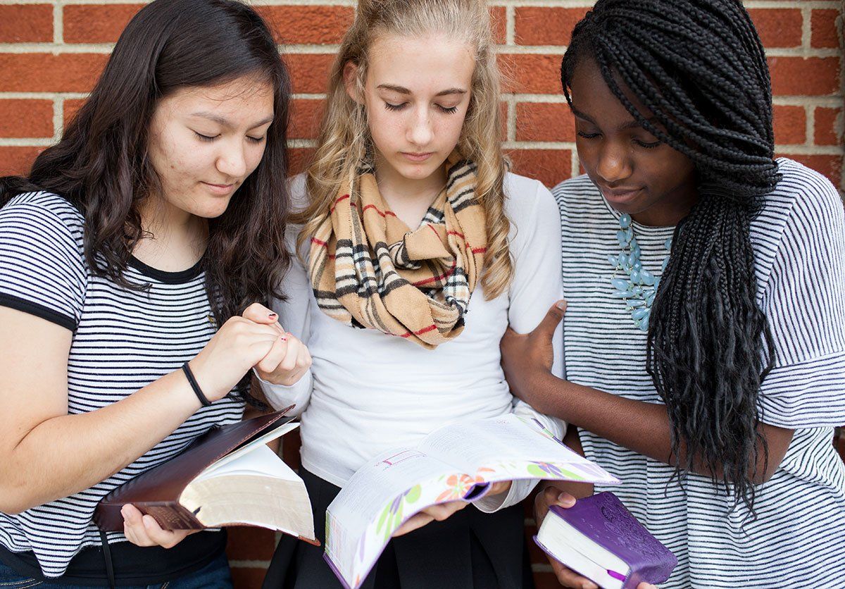 Three students stand against a brick wall, looking down at open books held in their hands.