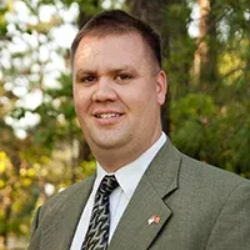 A smiling person in a green suit jacket, white collared shirt, and patterned tie, posed outdoors against a blurred forest.