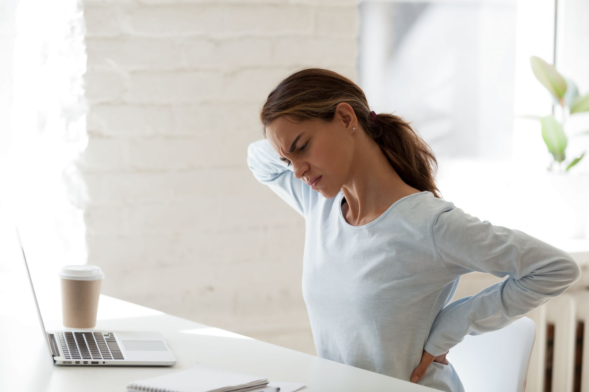 Woman sitting at a desk, holding her lower back beside a laptop and coffee, looking uncomfortable