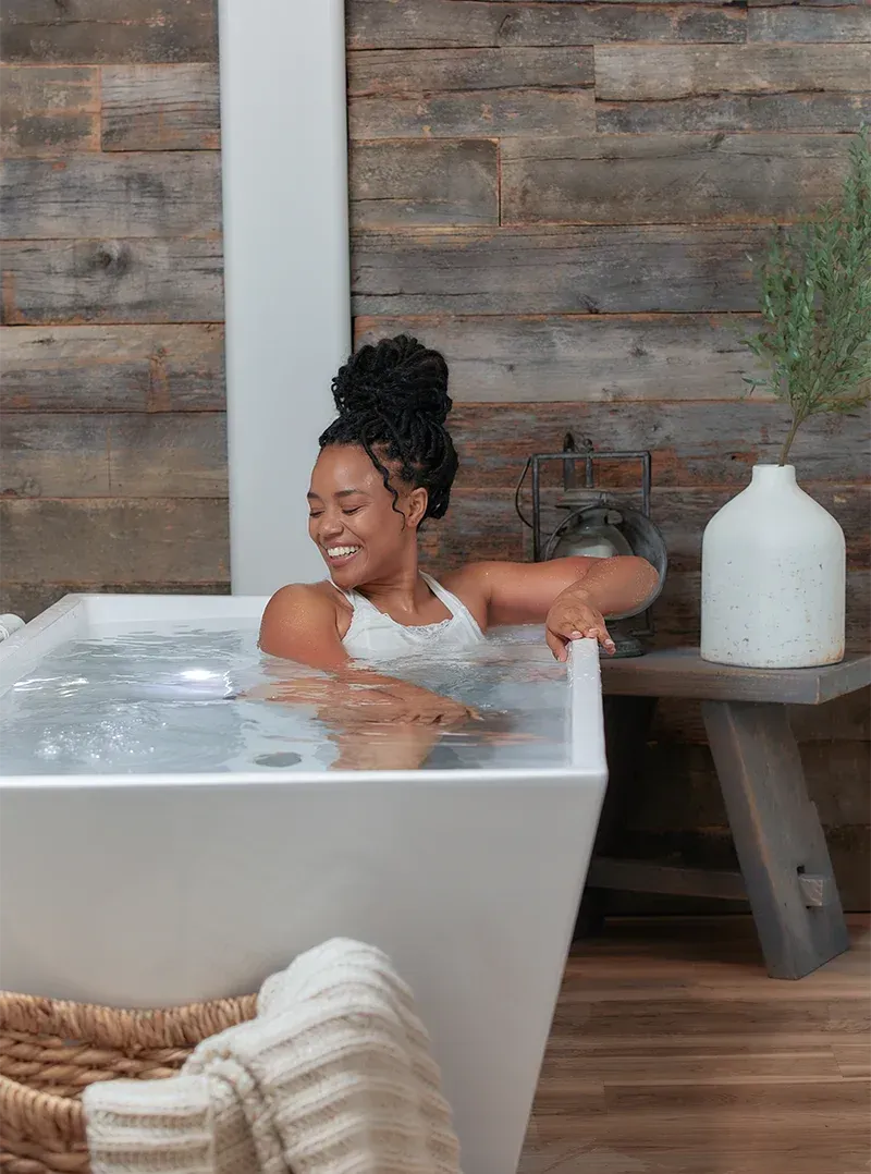 Person relaxing in a white bathtub with water, beside a wooden wall and decor in a cozy bathroom