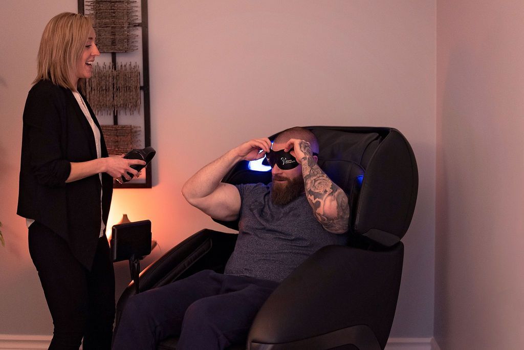 Person in a recliner using an eye mask while another person stands nearby in a room.