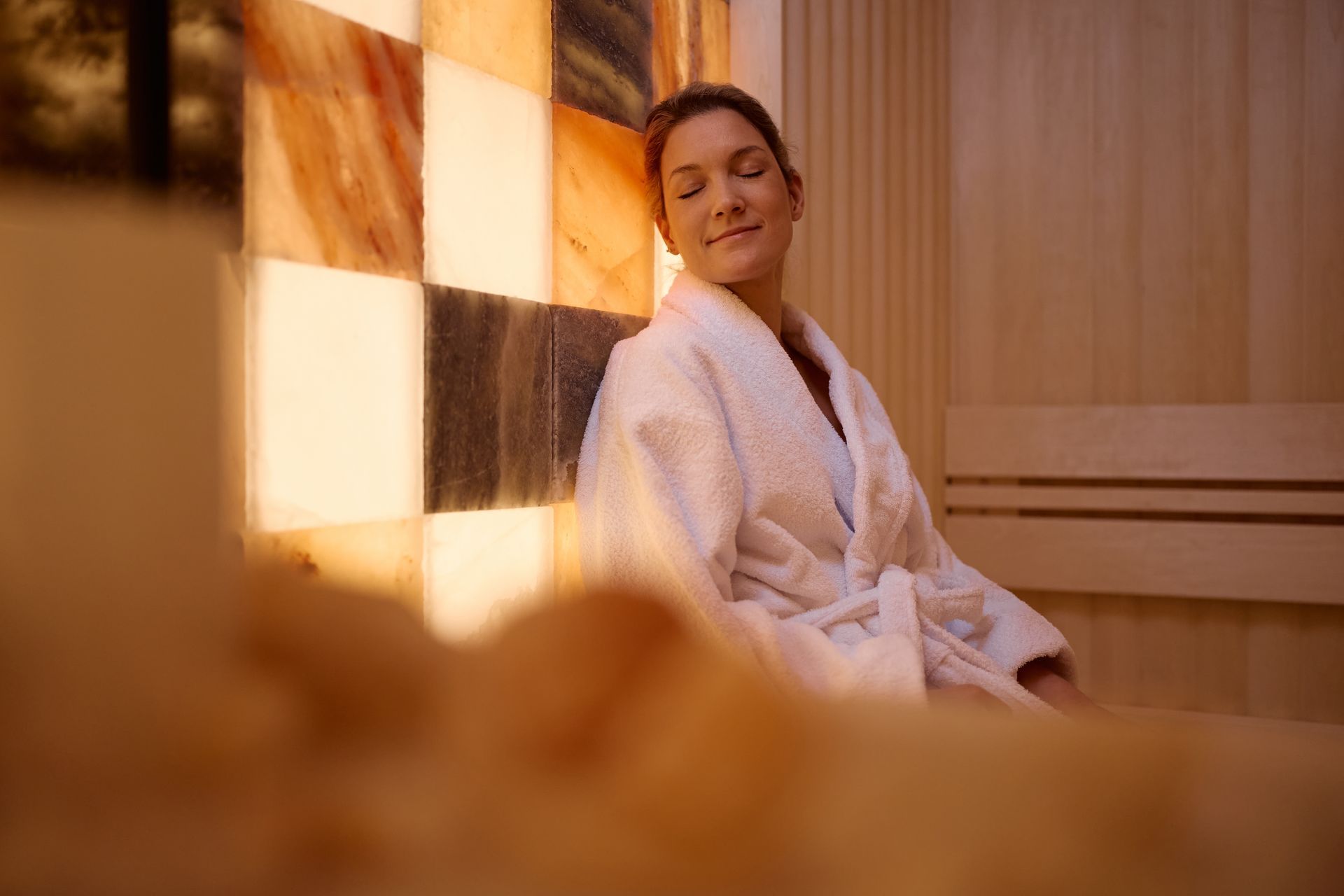 Person in a white robe relaxing in a warm spa sauna with checkered light on the wall.