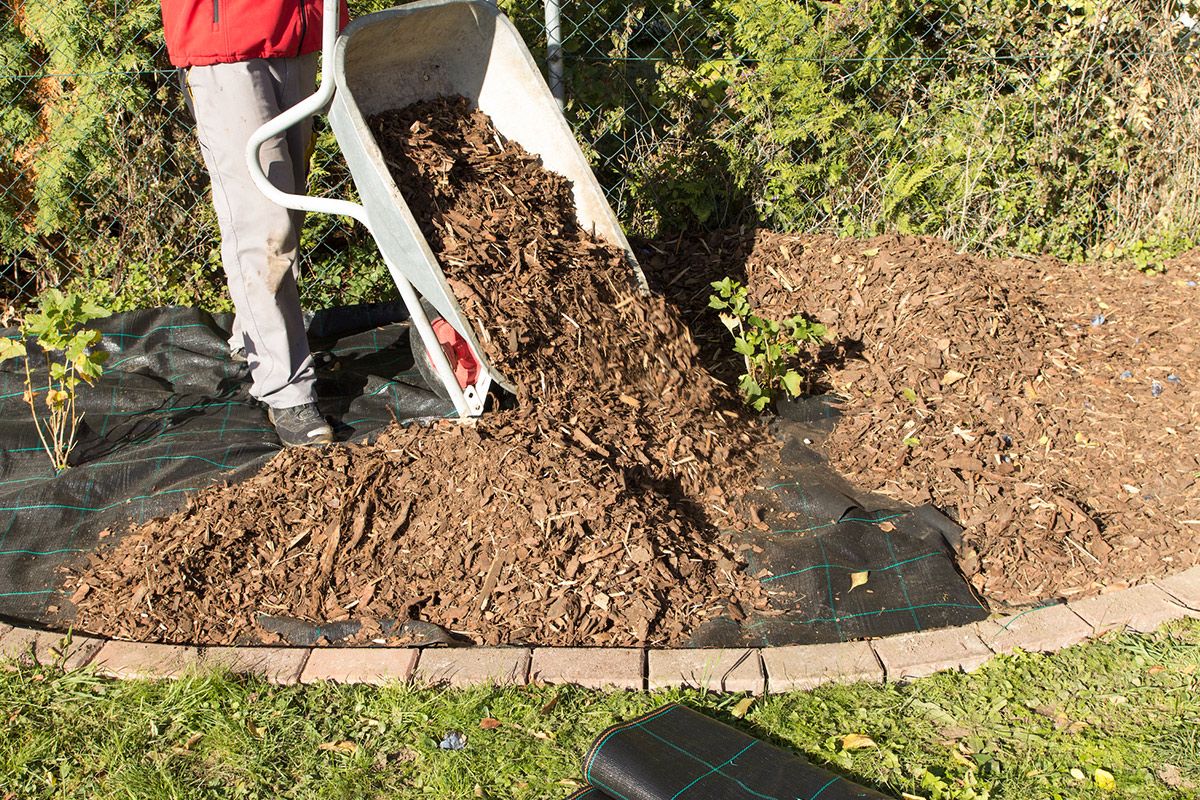 Person using a wheelbarrow to pour wood mulch onto a garden bed covered with black weed fabric, bordered by stone and grass.