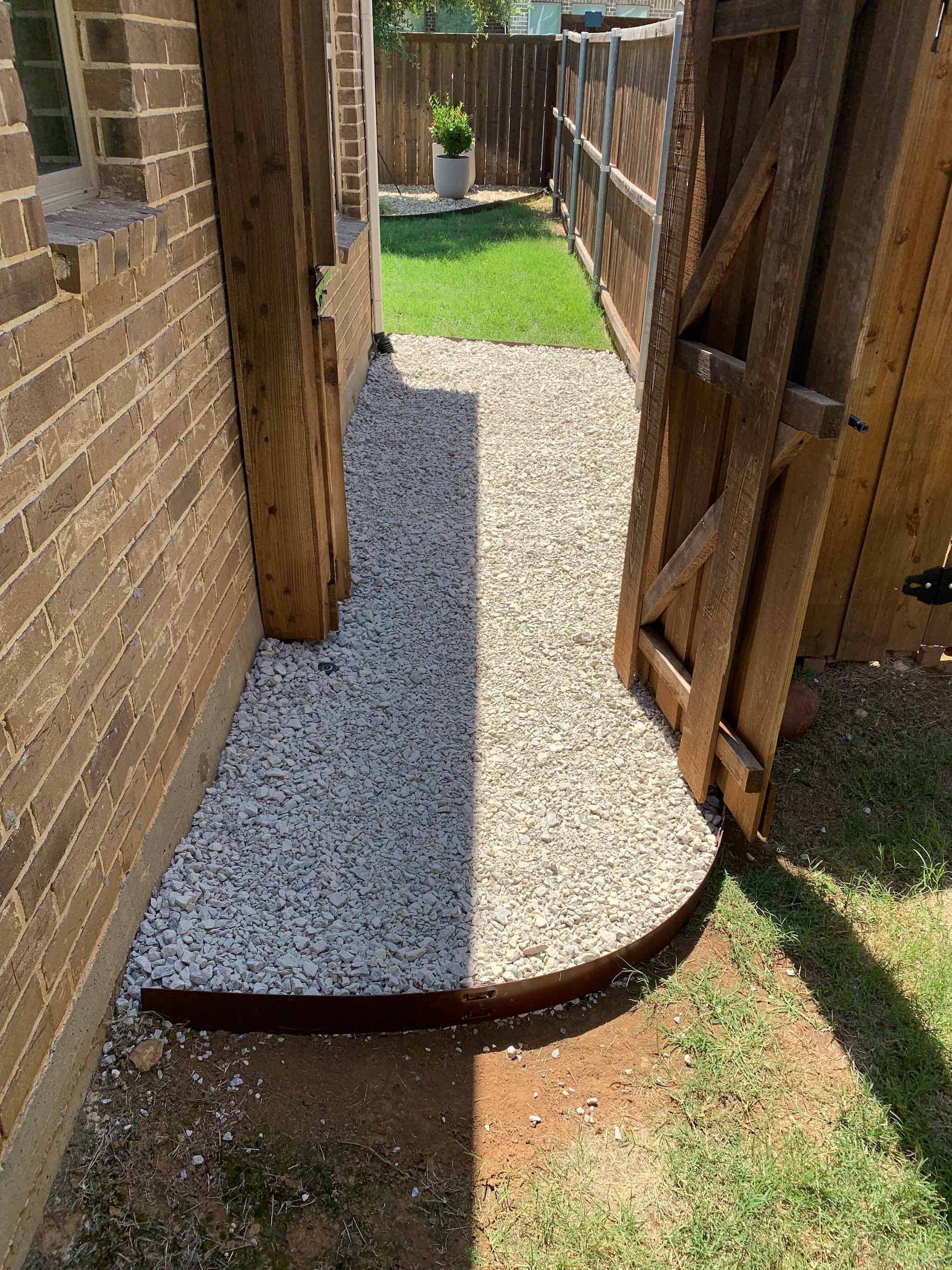 A narrow gravel path between a brick wall and wooden fence, leading to a gate and grassy area.