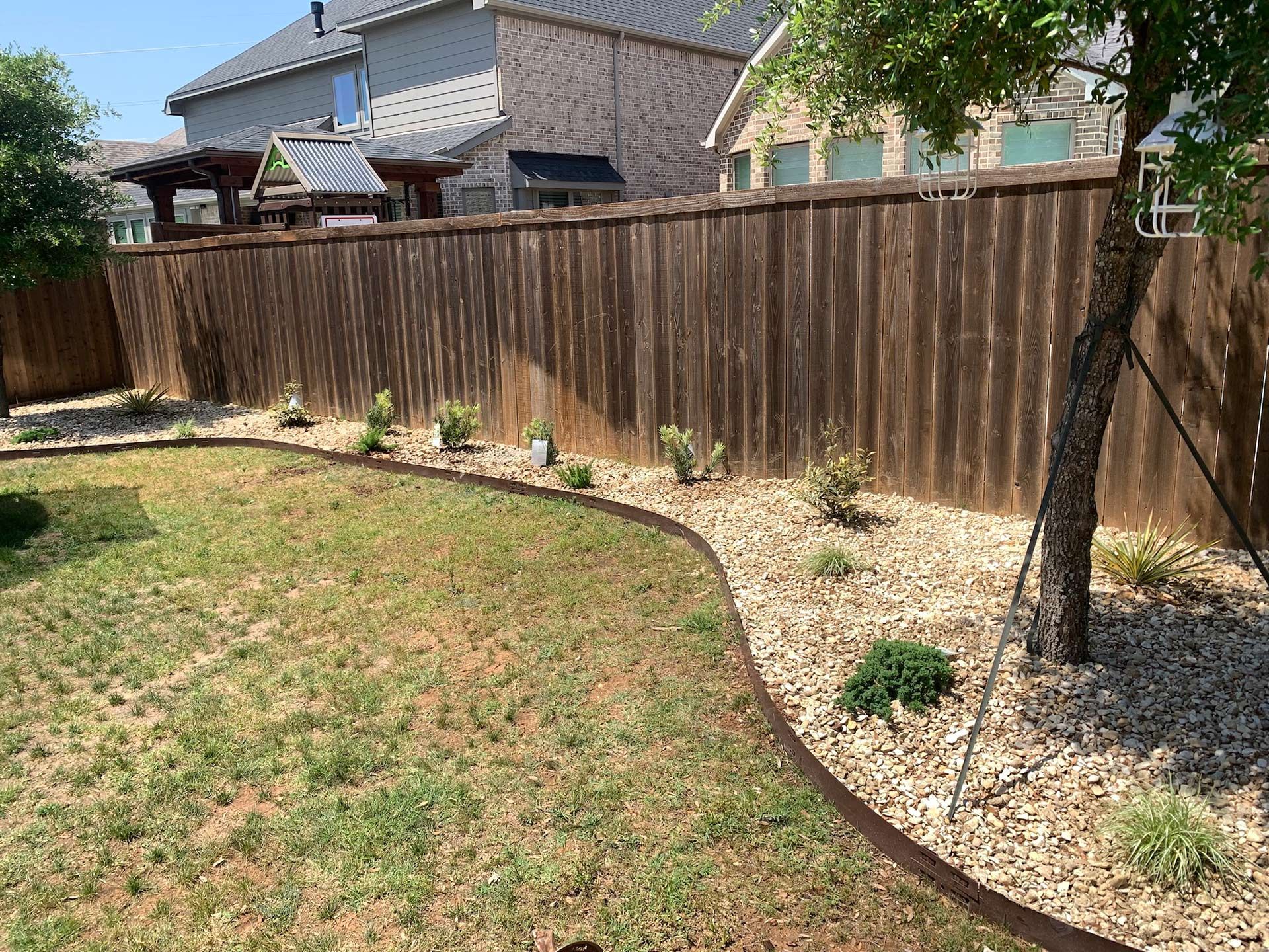 Backyard with wooden fence, brown ground cover, green grass, and small plants.