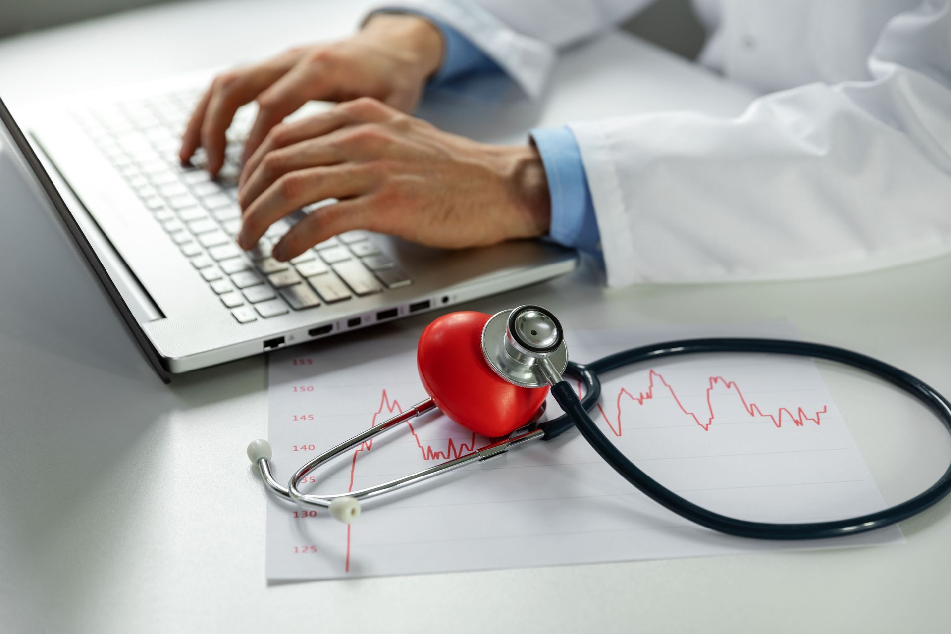 Médico digitando em um laptop com estetoscópio e modelo de coração vermelho em uma folha de eletrocardiograma.
