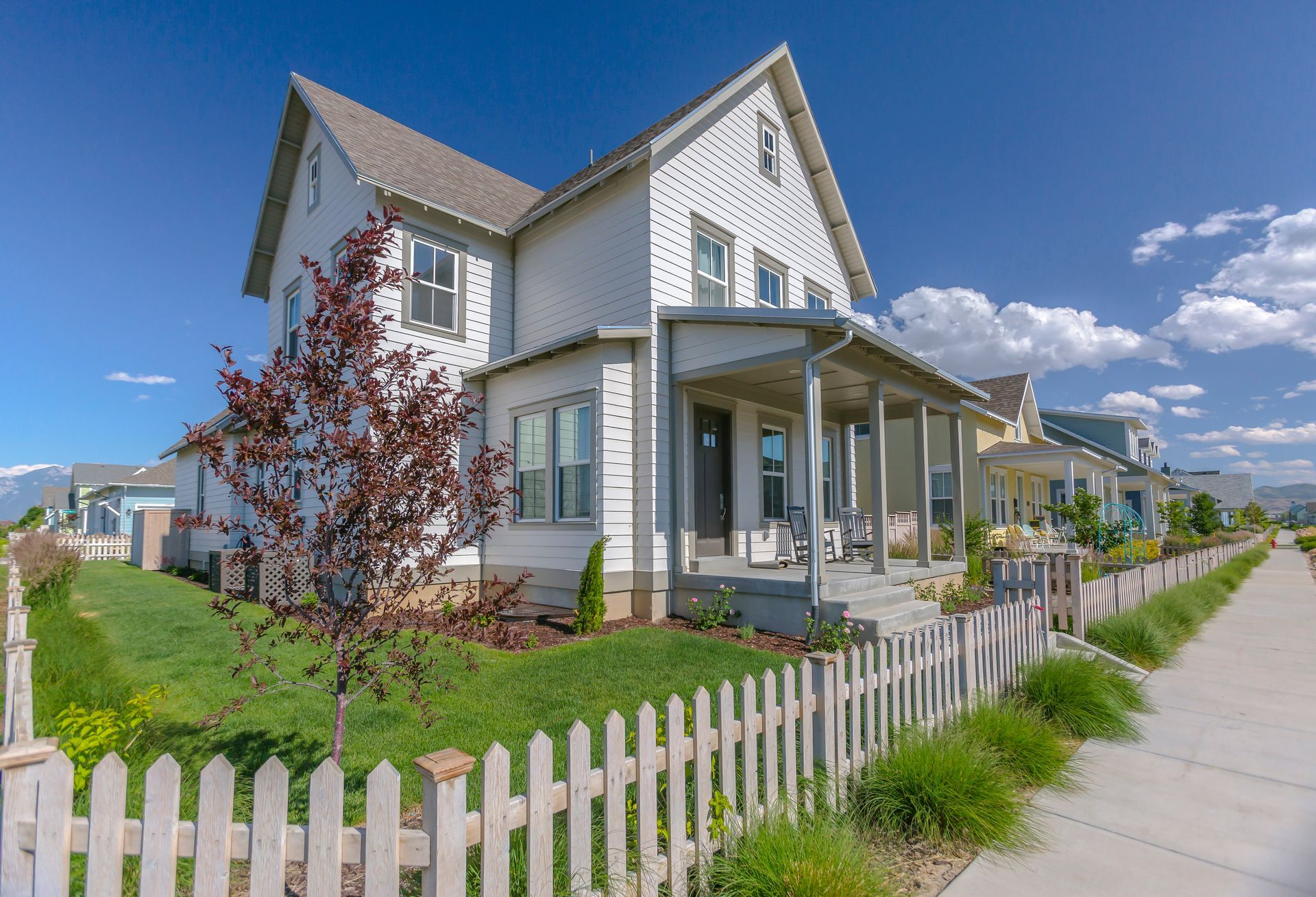 White two-story suburban house with a front porch, picket fence, and sidewalk under a blue sky