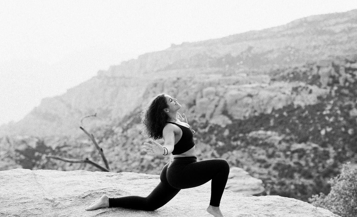 Woman in athletic wear doing yoga on a rocky cliff, mountain backdrop.