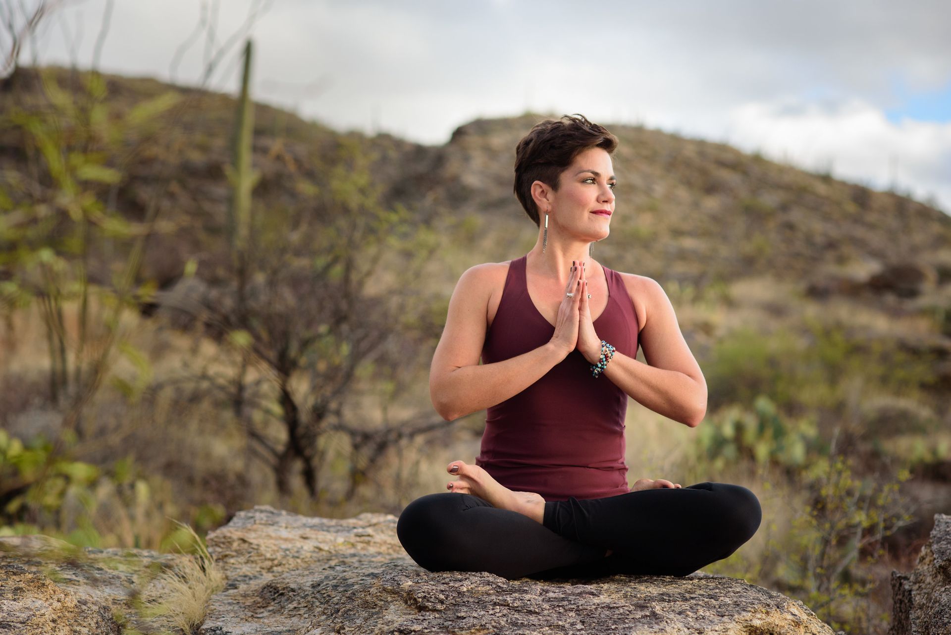 Woman in yoga pose outdoors, hands in prayer, looking right. Mountain backdrop.