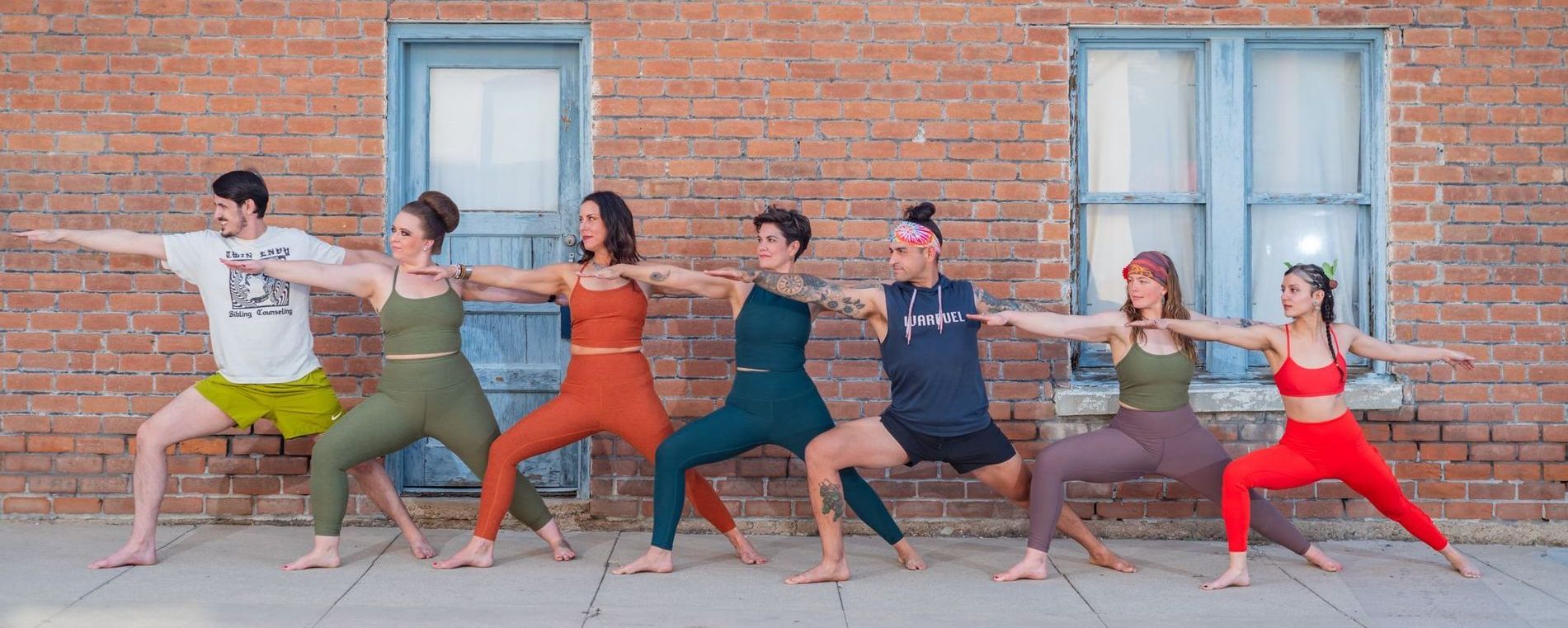 Seven people doing yoga poses in front of a brick building. Each wearing different colored workout clothes. Arms extended.