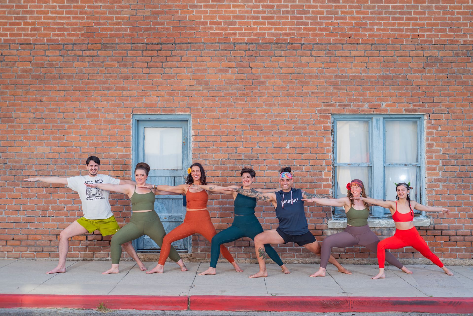Group of people in fitness poses in front of a brick wall, wearing workout clothes in various colors.