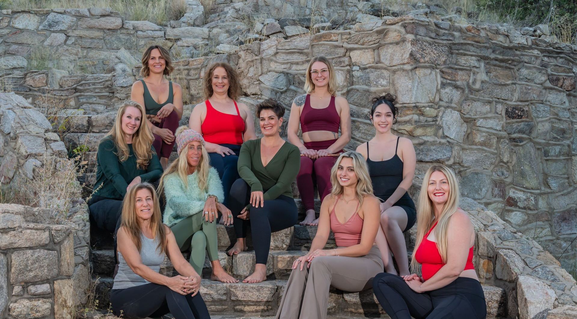 Group of women sitting on stone steps, smiling. Various athletic outfits. Outdoor setting.