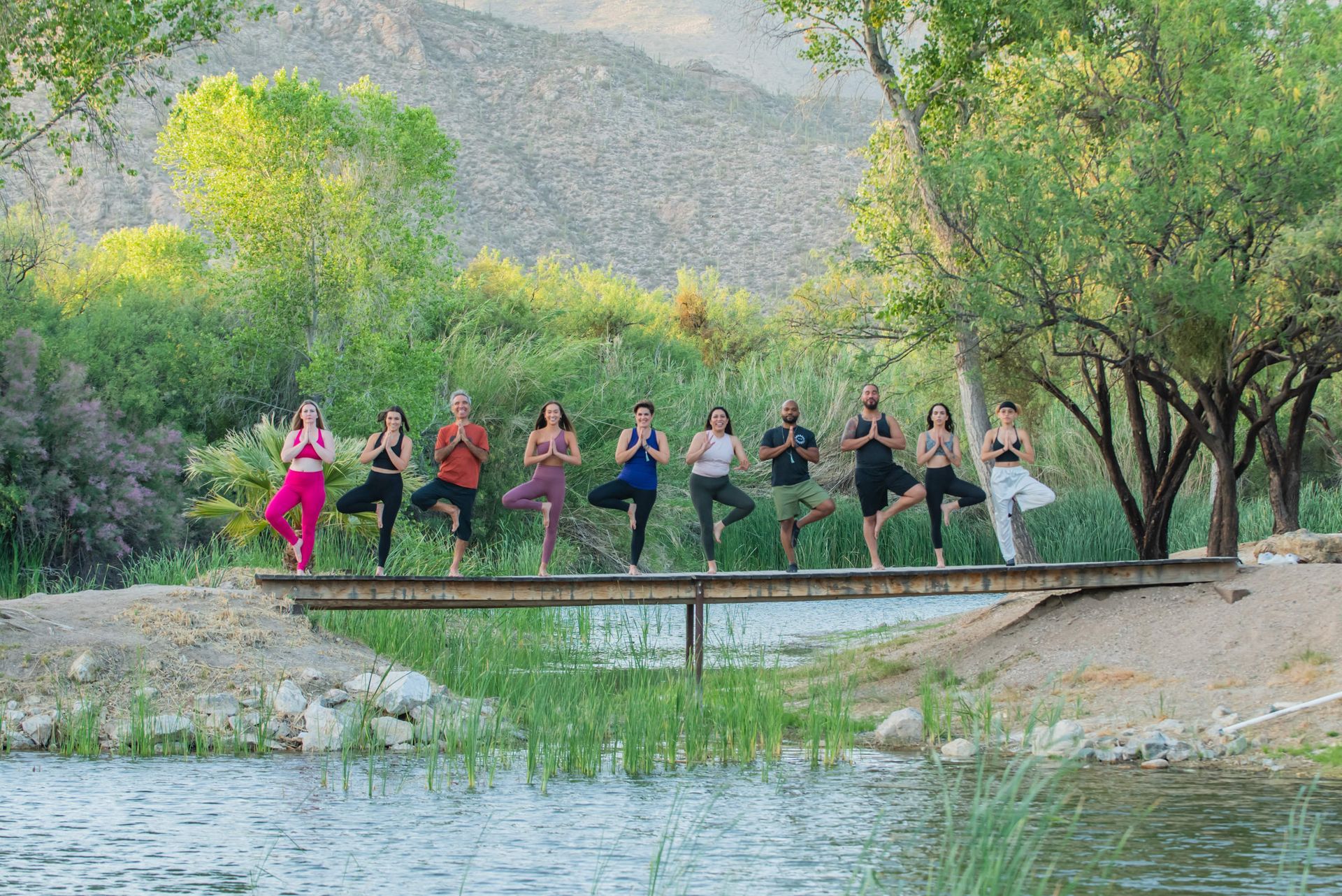 People practicing yoga on a wooden bridge over a pond, surrounded by nature.