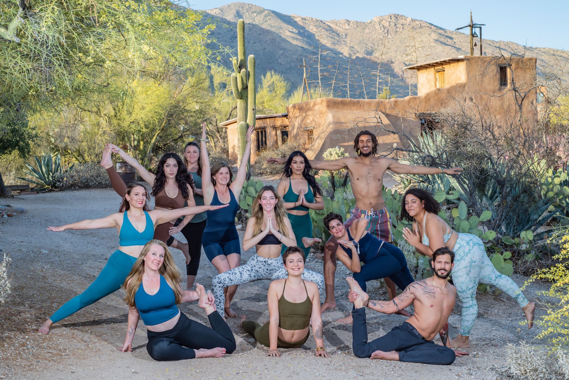 Group of people doing yoga poses outdoors near a desert-like structure, mountains, and greenery.
