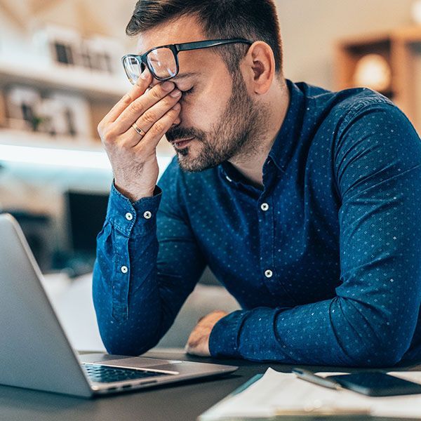 Man in blue shirt rubbing his eyes while working on a laptop at a desk