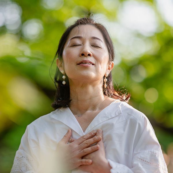 Woman with eyes closed, hands over heart, standing outdoors in sunlight with blurred green background